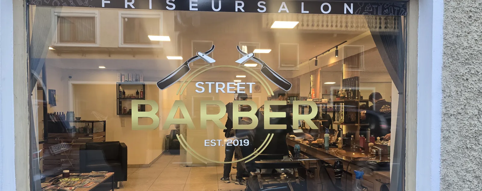 Schaufenster mit goldenem Street Barber Logo „Est. 2019“ und Blick in den Salon